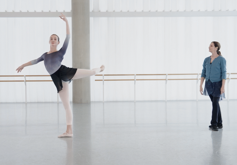 Ballet dancer in gray leotard performs an arabesque at the barre in a bright white studio, with one leg extended behind her and arm raised gracefully overhead. An instructor in a teal cardigan observes from across the room.