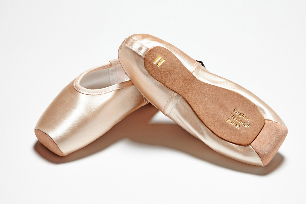 A pair of pink satin pointe ballet shoes positioned on a light gray surface, one lying on its side showing the sole branded 'English National Ballet' and the other standing upright.