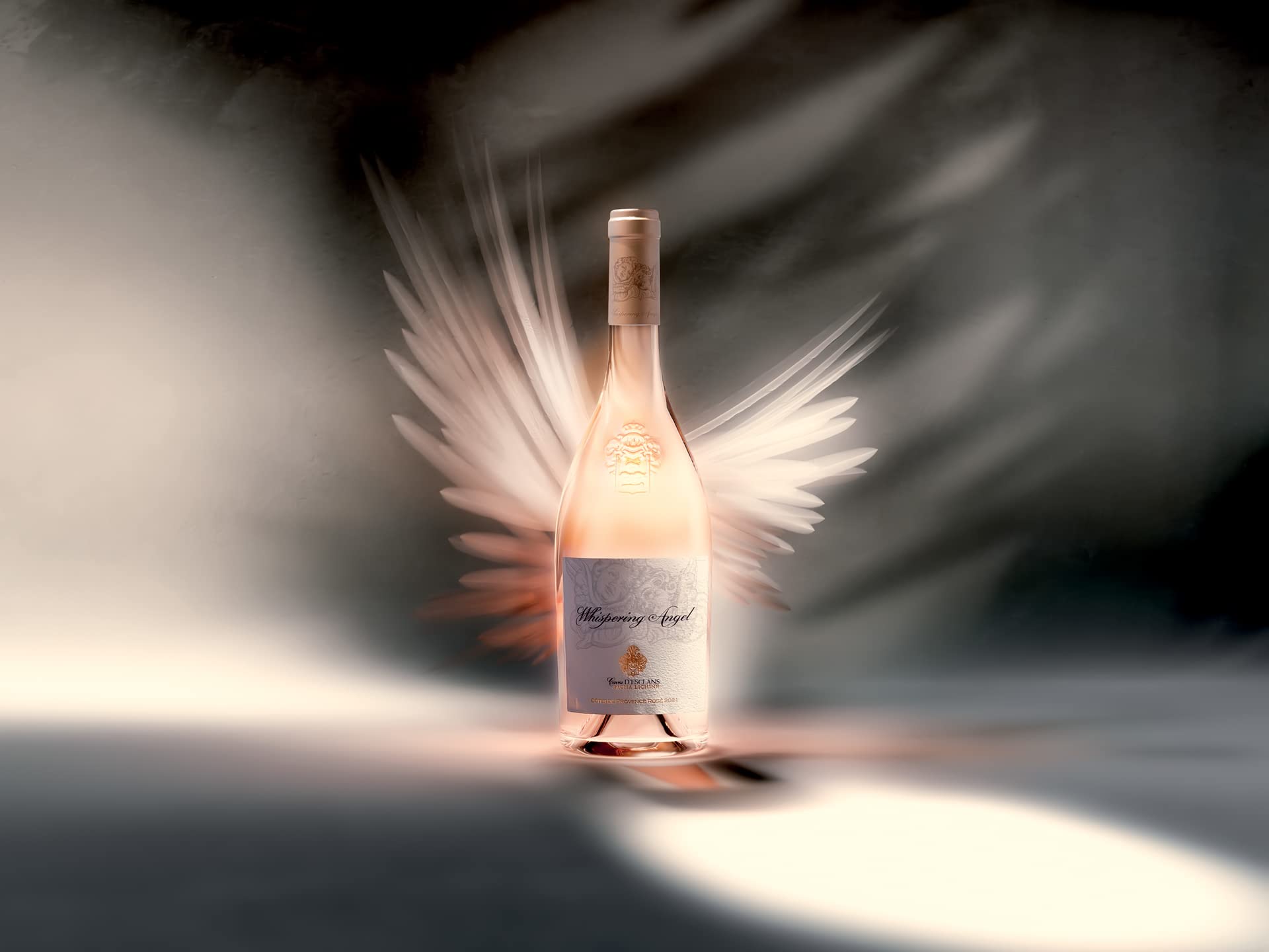 A bottle of Whispering Angel rosé wine stands upright against a soft, atmospheric background. Ethereal, feather-like white wings appear to glow behind the bottle.