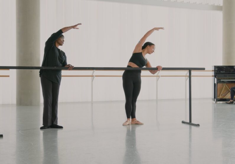 Two dancers stand at a ballet barre in a bright studio, both leaning to the side with one arm curved overhead in a stretching position. A pianist sits at a piano in the background, watching them.