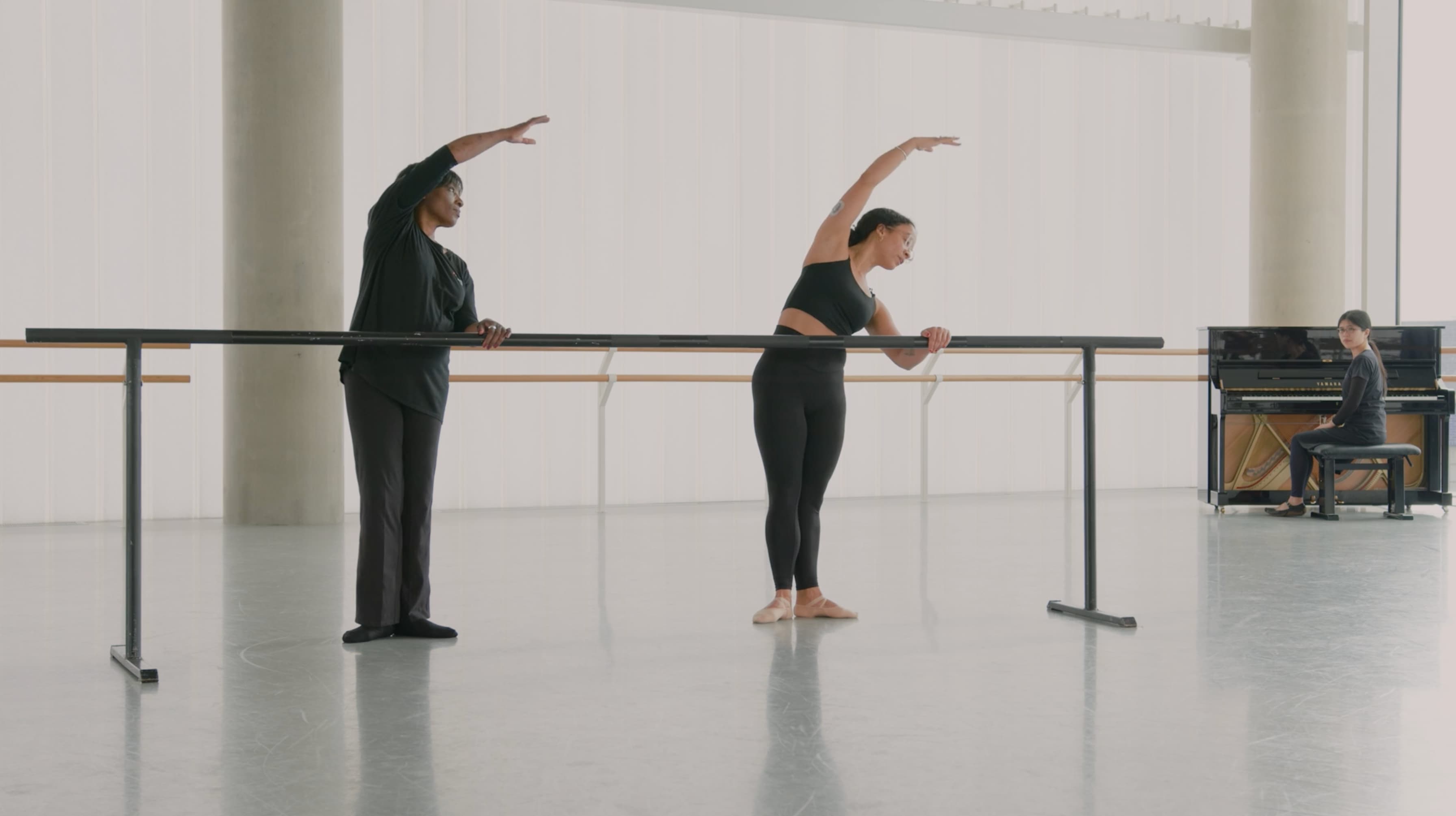 Two dancers stand at a ballet barre in a bright studio, both leaning to the side with one arm curved overhead in a stretching position. A pianist sits at a piano in the background, watching them.