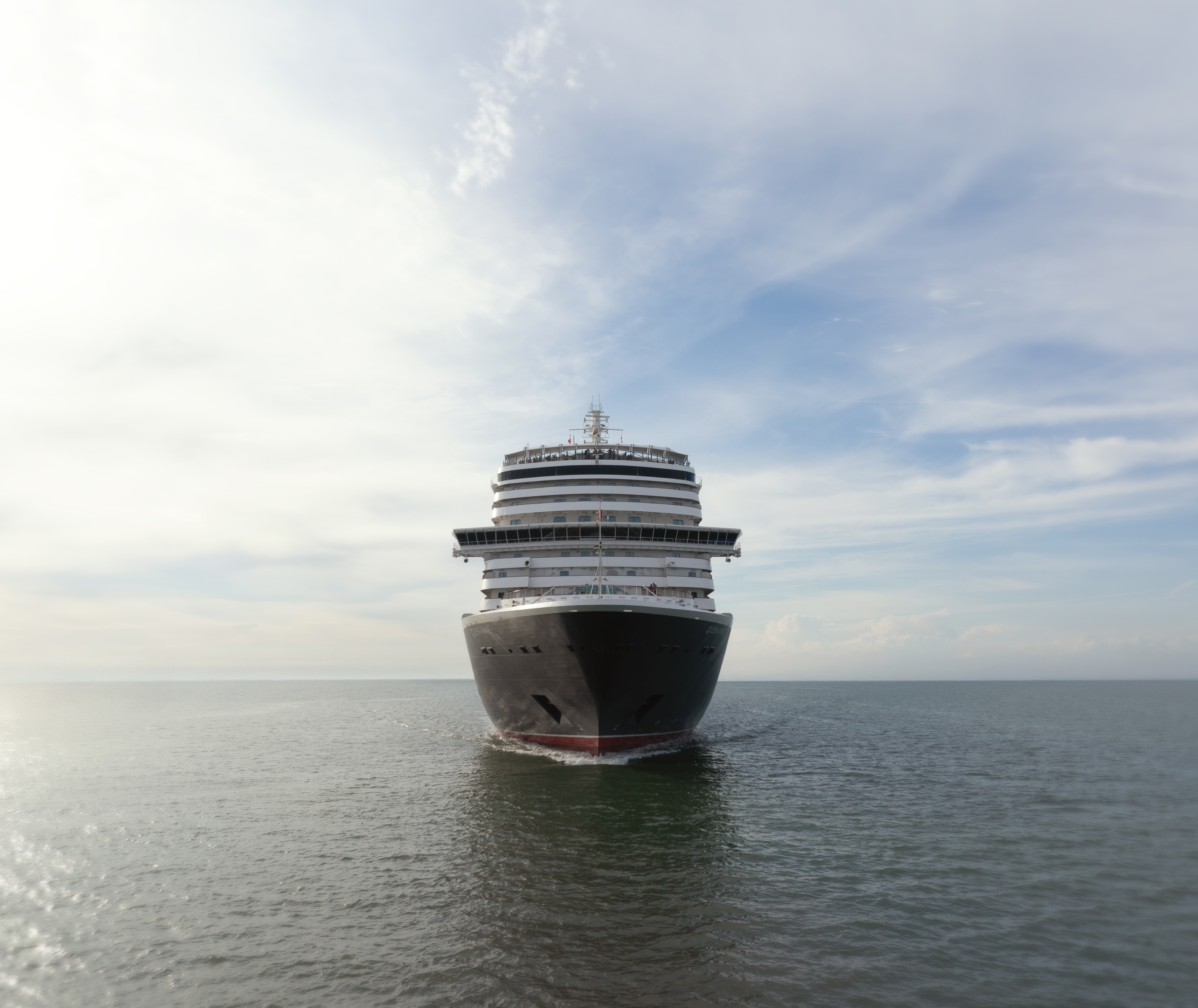 A large cruise ship sails forward on calm open water, photographed head-on under a bright sky with scattered clouds.
