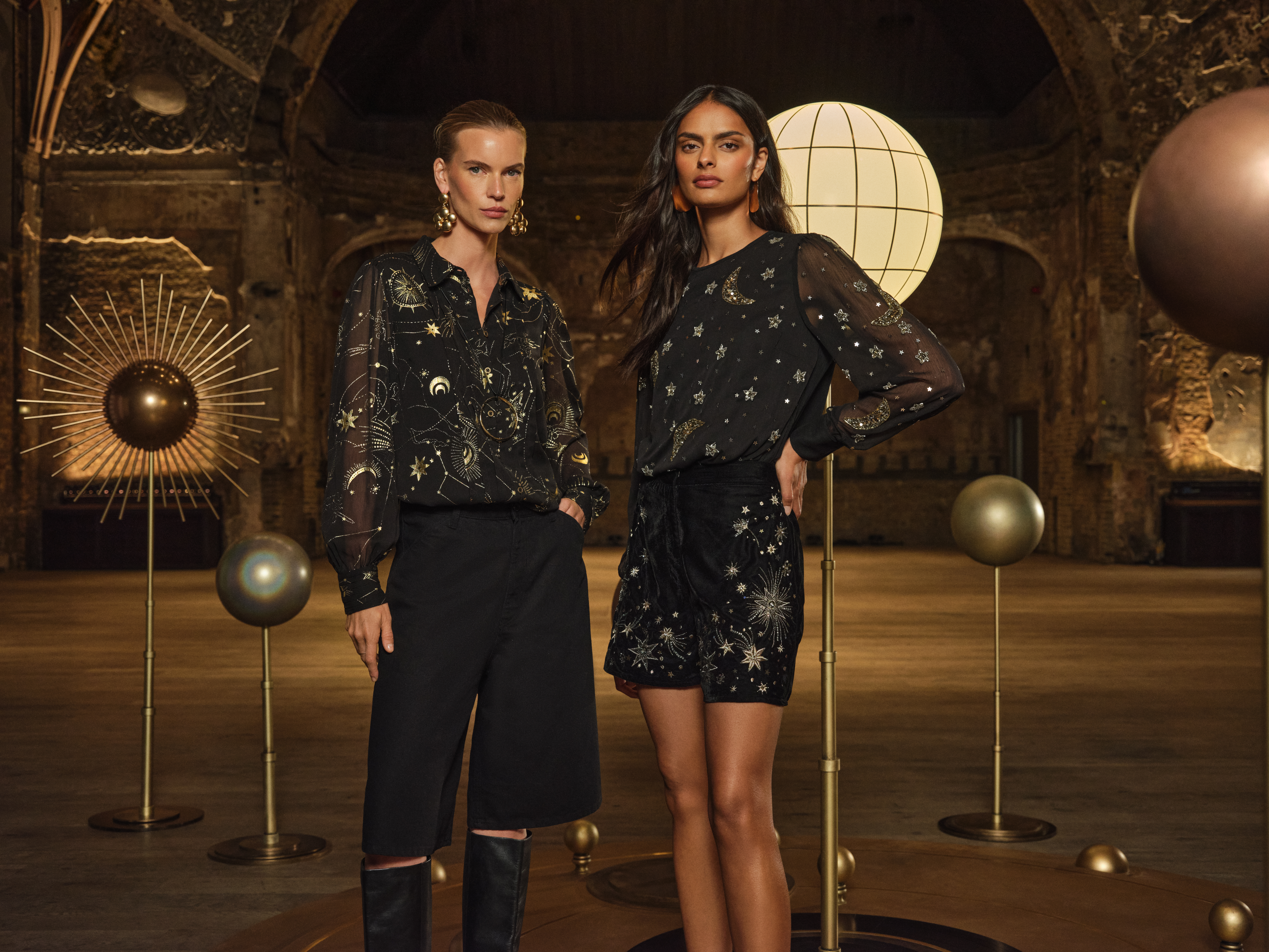 Two women stand in an ornate, dimly lit hall decorated with celestial-themed sculptures. Both wear black outfits with gold star, moon, and constellation embellishments.
