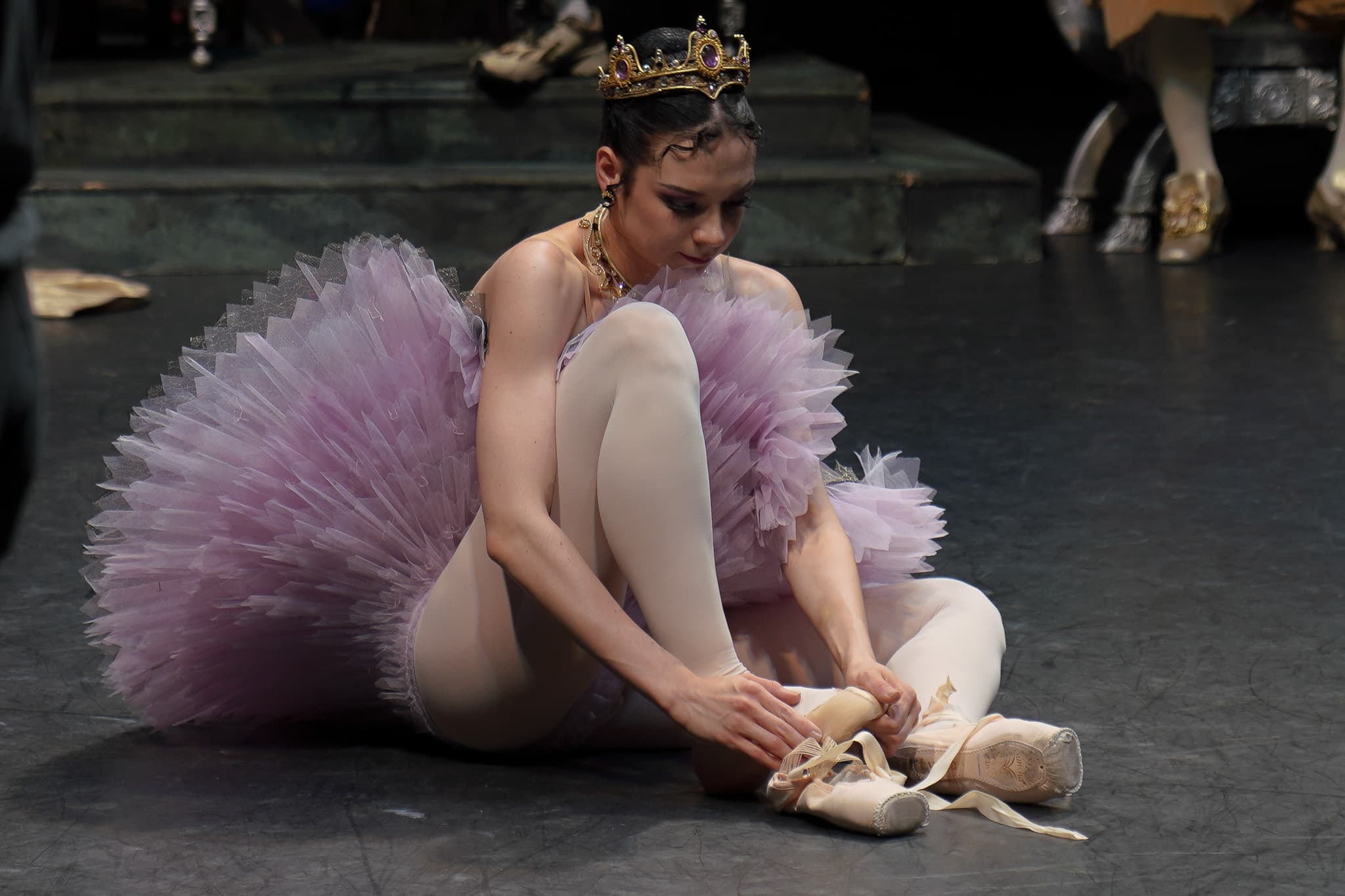A ballet dancer wearing a lavender tutu, pink tights, and a jewelled crown sits on stage tying the ribbons of her pointe shoes, preparing for a performance.