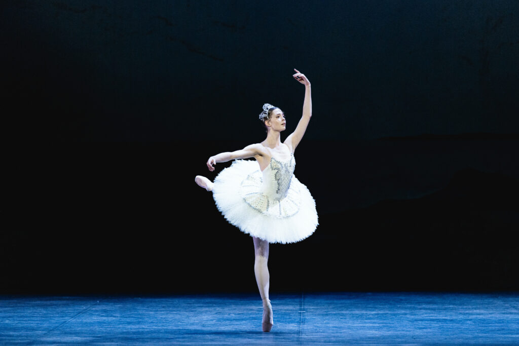 A ballet dancer in a white classical tutu balances en pointe in an elegant arabesque on a dimly lit stage, one arm extended to the side and the other raised overhead, with a dark backdrop behind her.