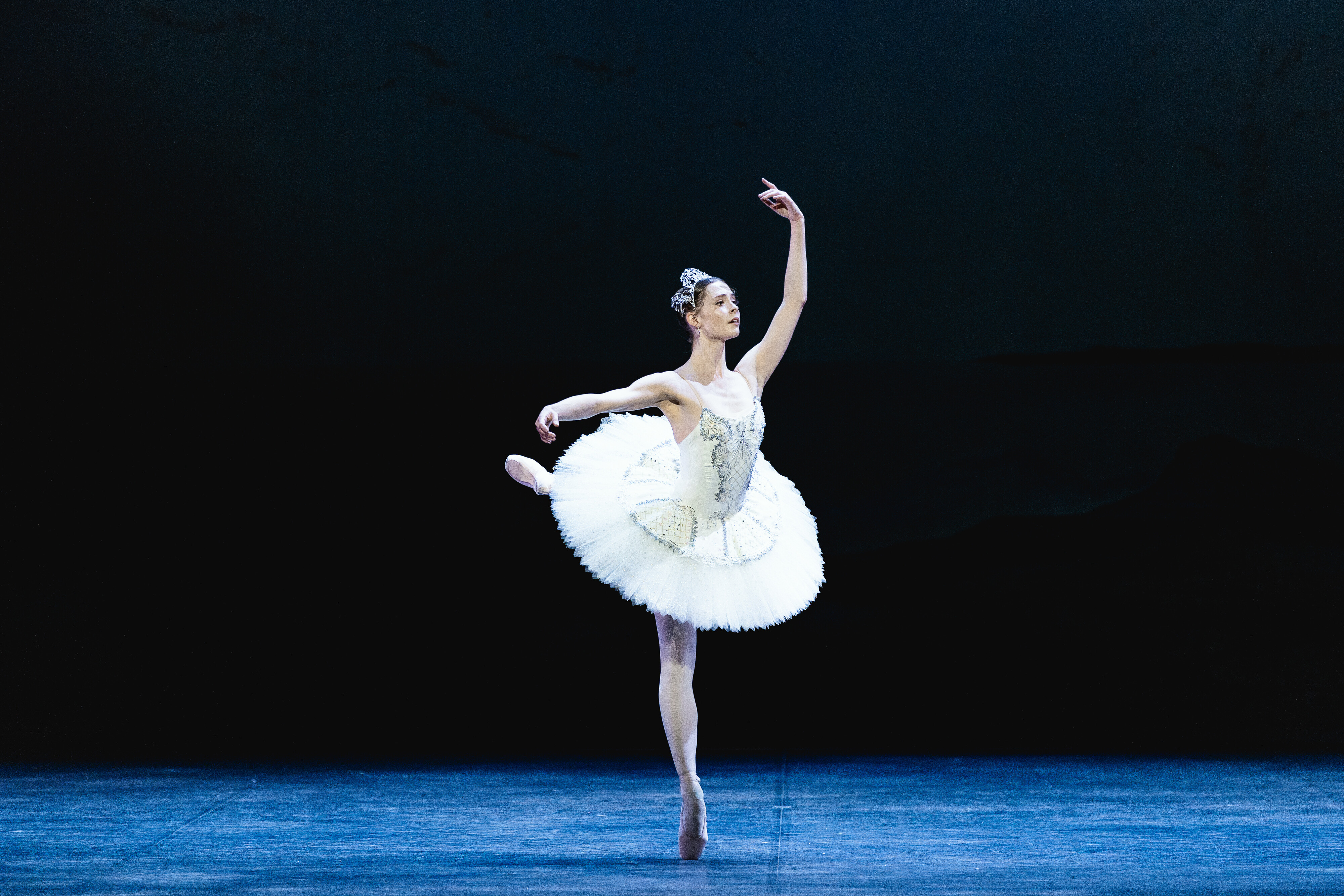A ballet dancer in a white classical tutu balances en pointe in an elegant arabesque on a dimly lit stage, one arm extended to the side and the other raised overhead, with a dark backdrop behind her.