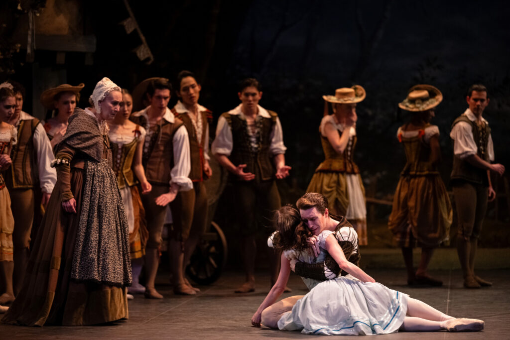 A ballet performance showing two dancers in the foreground - a male dancer kneeling behind and embracing a female dancer in a white dress who is seated on the stage floor. Behind them, a group of ensemble dancers in period costumes watch the scene.
