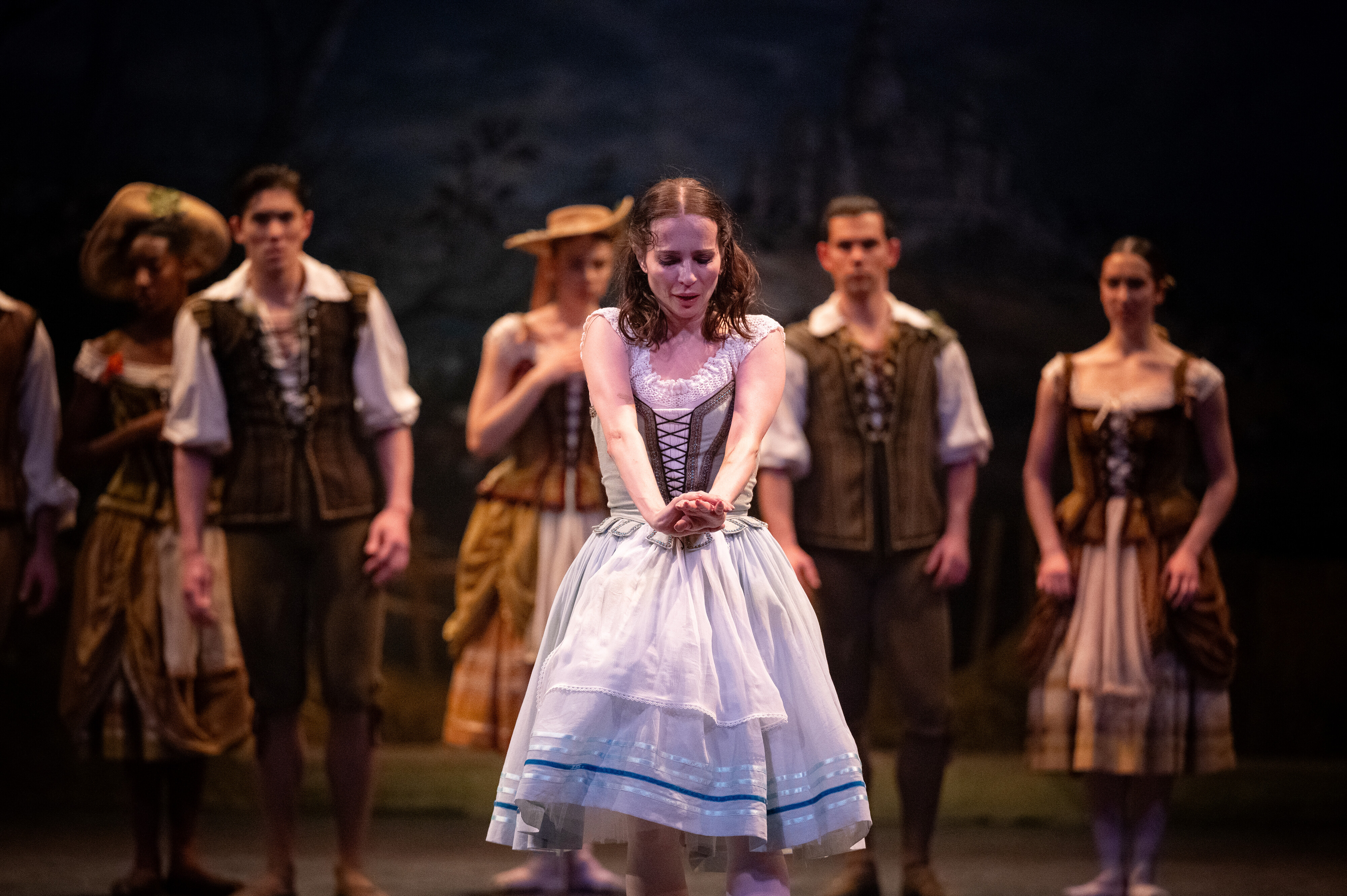 A female ballet dancer in a white peasant-style dress and corset stands center stage with her arms crossed and extended downward, performing an expressive moment from Giselle. Several other dancers in rustic village costumes stand behind her, watching intently.