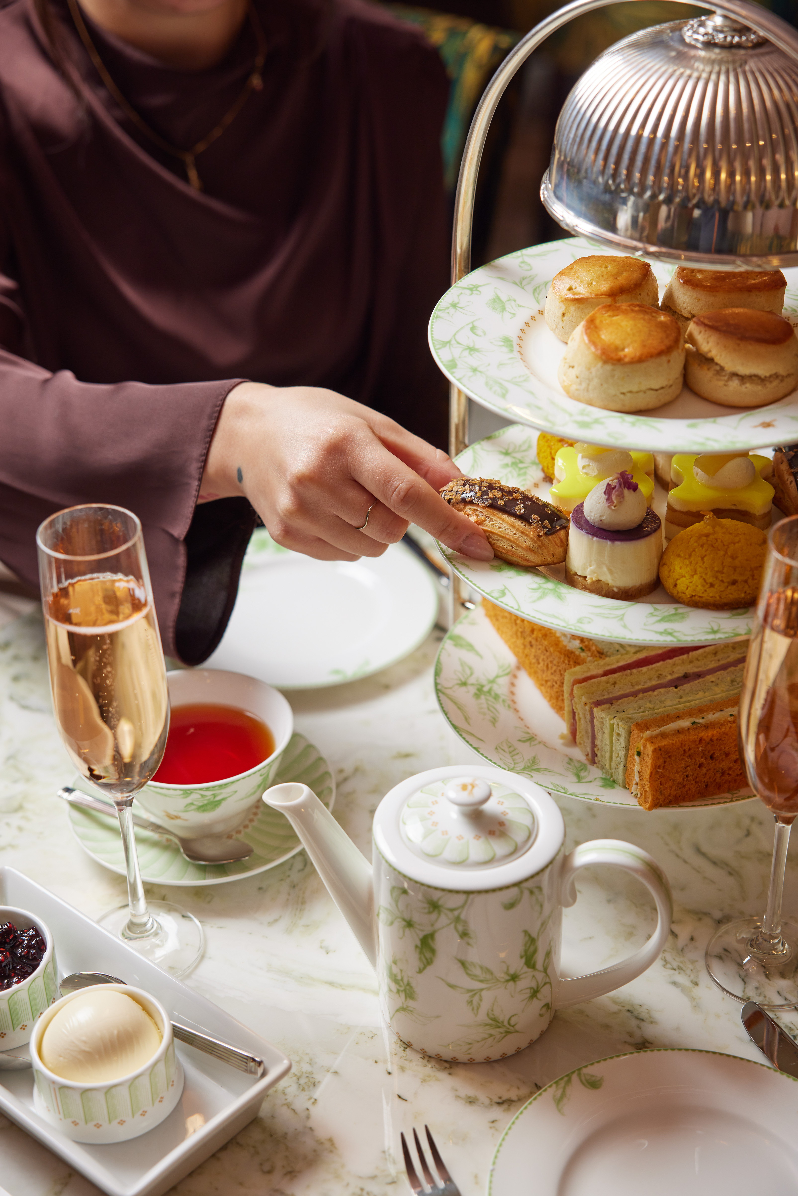 A close-up of an elegant afternoon tea setting, showing a person reaching for a chocolate-topped pastry from a tiered stand filled with scones, cakes, and finger sandwiches.