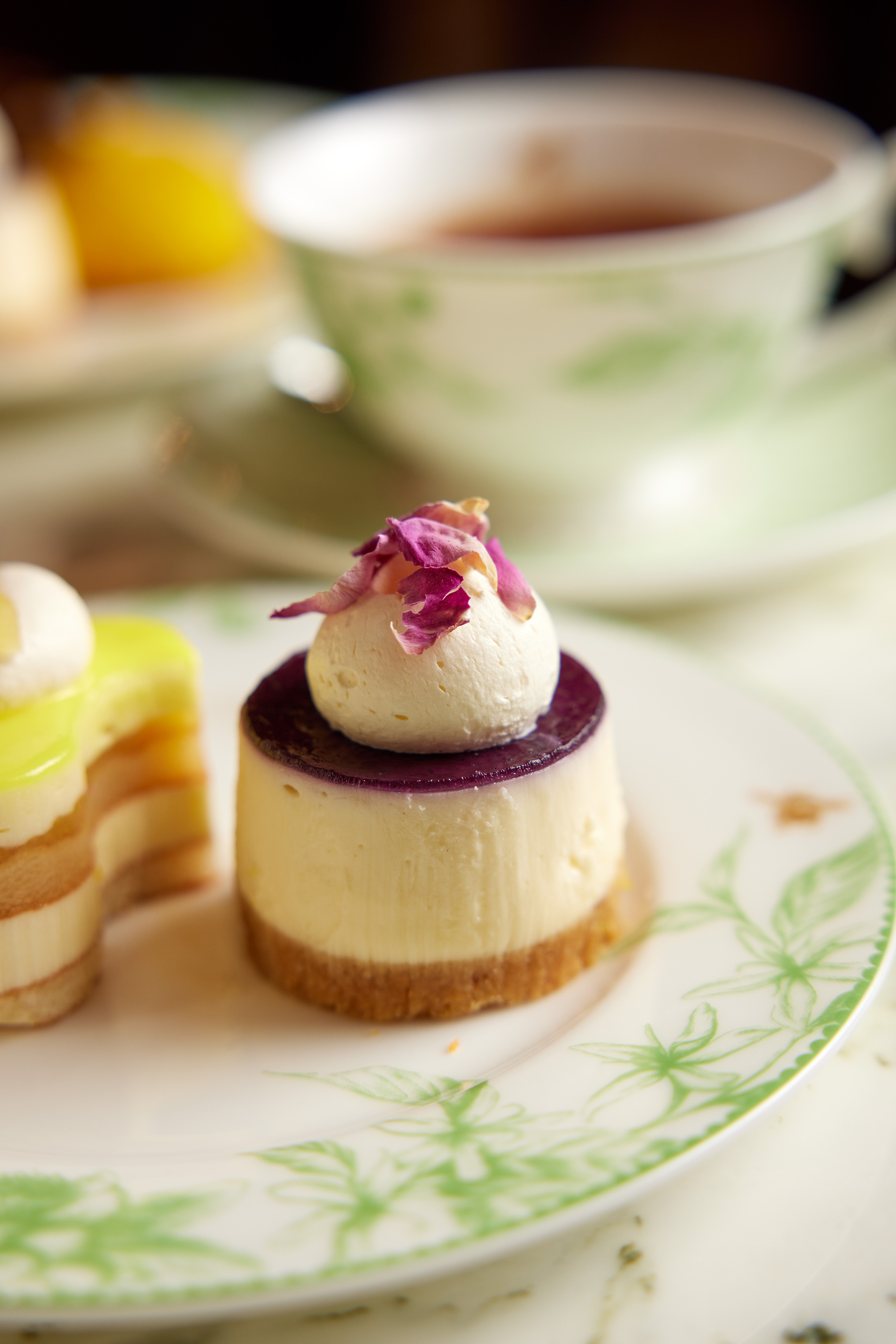 Mini cheesecake with a biscuit base, purple fruit topping, and pink flower petals, served on a plate with tea in the background.