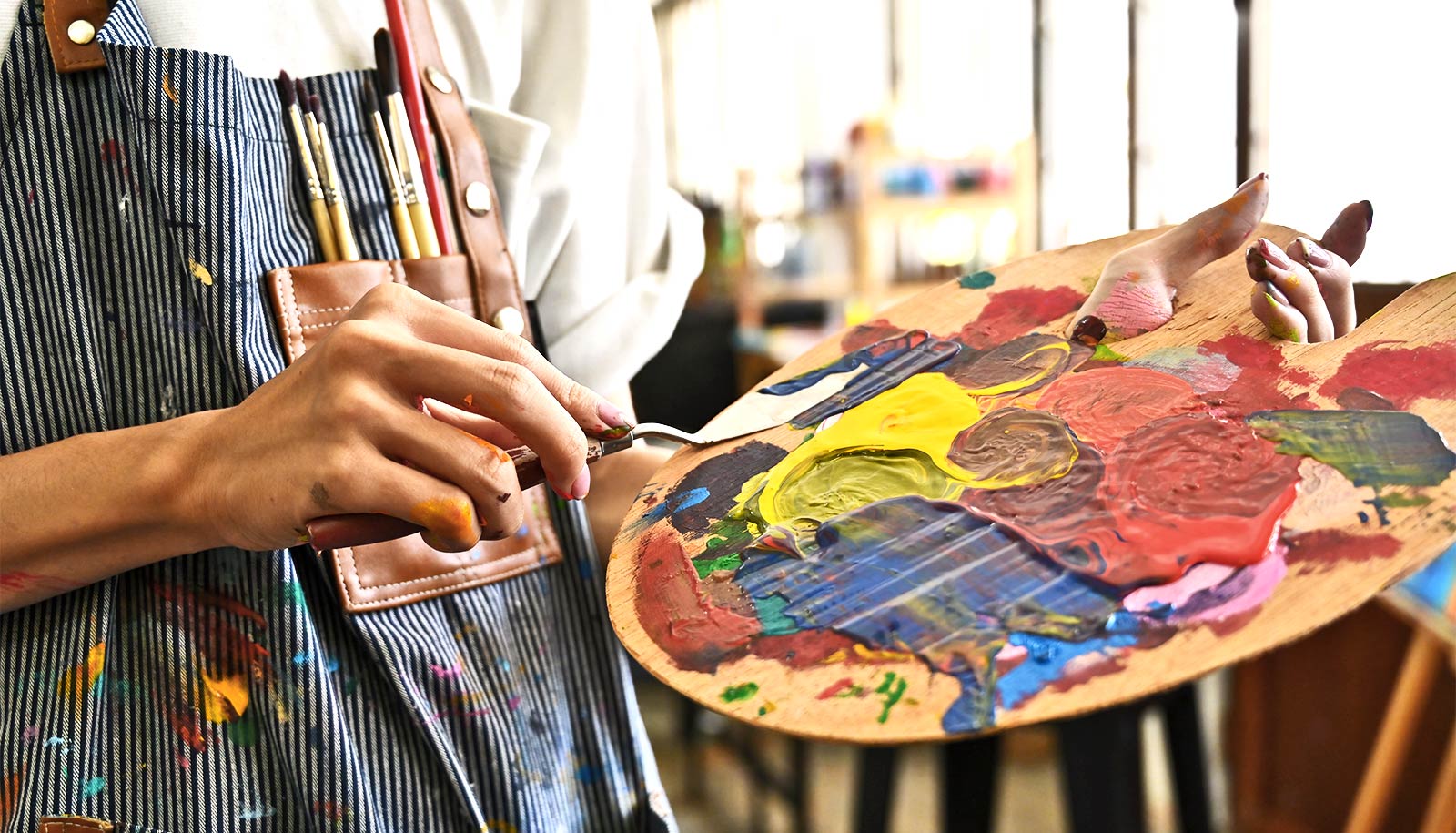 Close-up of an artist holding a wooden paint palette covered in mixed, colourful paints while wearing a paint-splattered apron, with brushes tucked into the pocket and a studio setting softly blurred in the background.