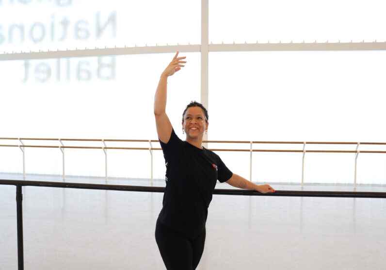 A dancer stands at a ballet barre in a bright studio, smiling while holding one arm curved above her head and the other resting on the barre.