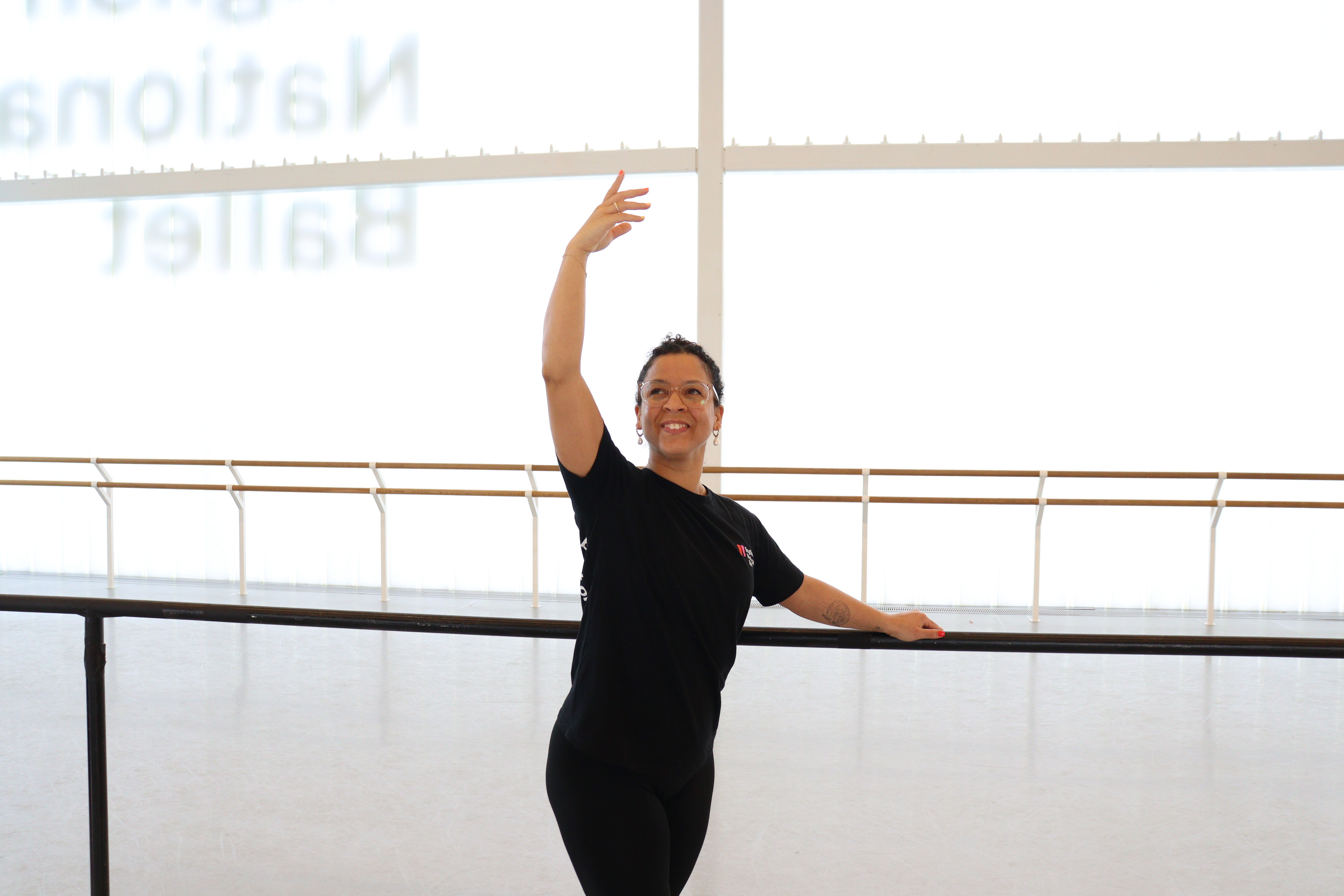A dancer stands at a ballet barre in a bright studio, smiling while holding one arm curved above her head and the other resting on the barre.