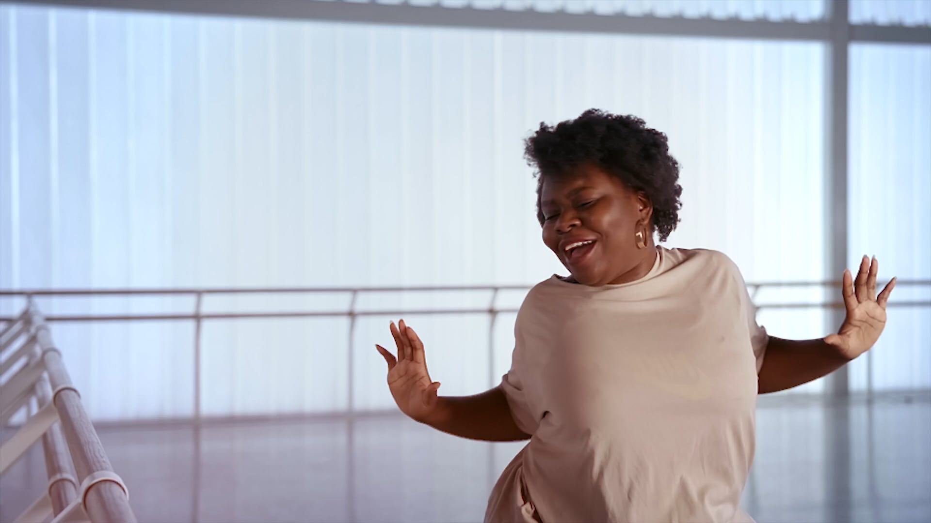 A person dancing joyfully in a bright studio, smiling with eyes closed and arms raised, standing near a ballet barre with large windows in the background.