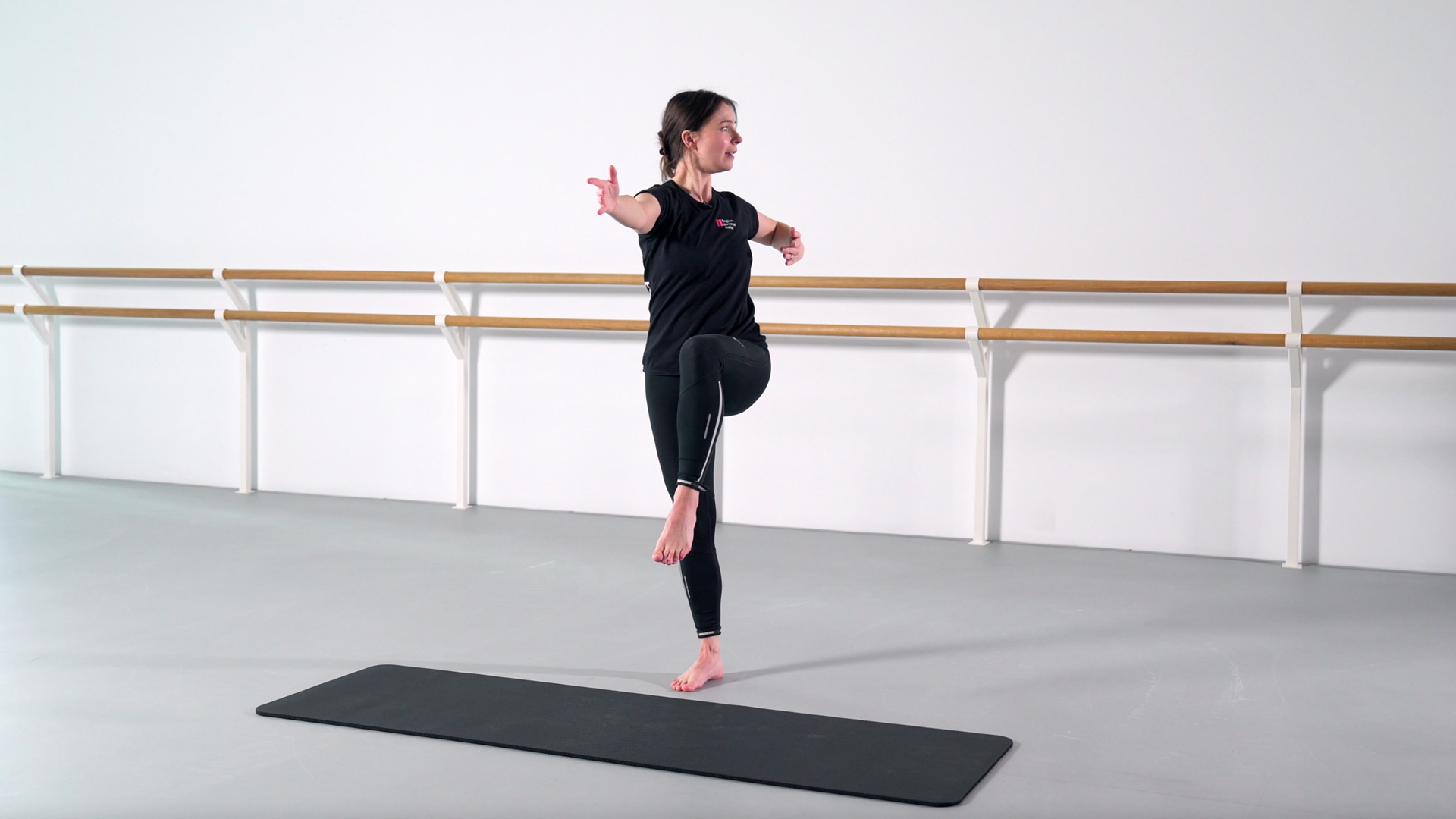 A woman practices a balance exercise in a dance studio, standing on one leg with the other knee lifted and arms extended, barefoot on a mat in front of ballet barres.