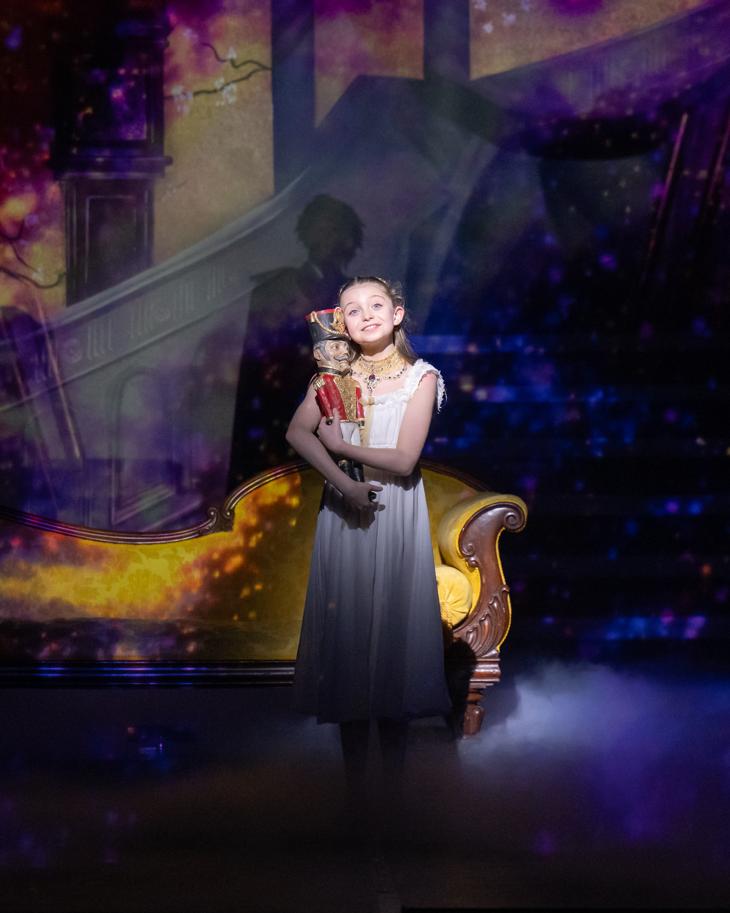 A young girl in a white nightdress stands onstage, smiling softly as she holds a toy nutcracker. She is illuminated by a warm spotlight. Behind her, a golden sofa and a backdrop of a grand staircase and colourful, sparkling projections create a magical, festive atmosphere.