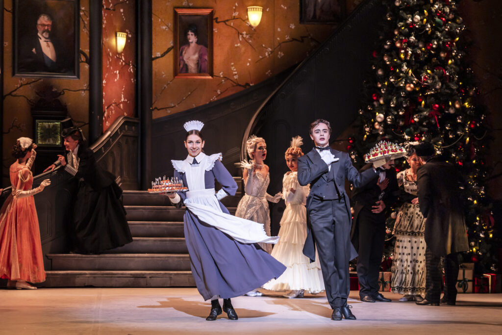 A festive stage scene from a ballet production of Nutcracker, showing a maid and a butler in period costumes presenting trays of miniature soldier figurines as they pose at the front of a grand Victorian-style room.