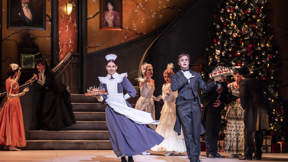 A festive stage scene from a ballet production of Nutcracker, showing a maid and a butler in period costumes presenting trays of miniature soldier figurines as they pose at the front of a grand Victorian-style room.