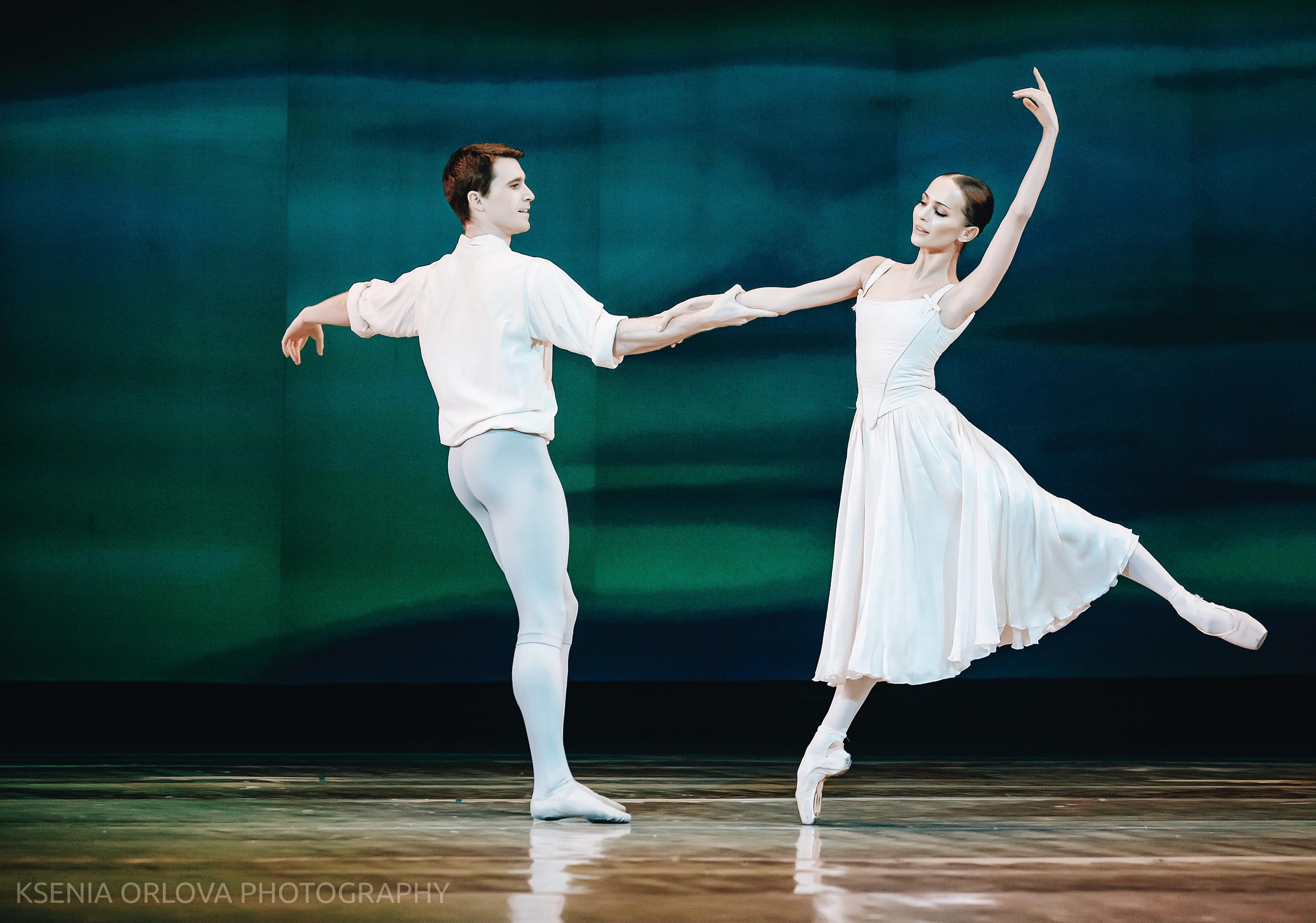 A ballet dancer in a white dress balances en pointe with one arm lifted gracefully overhead while holding hands with her partner. Her partner, dressed in white tights and a soft shirt, supports her as they face each other on stage.