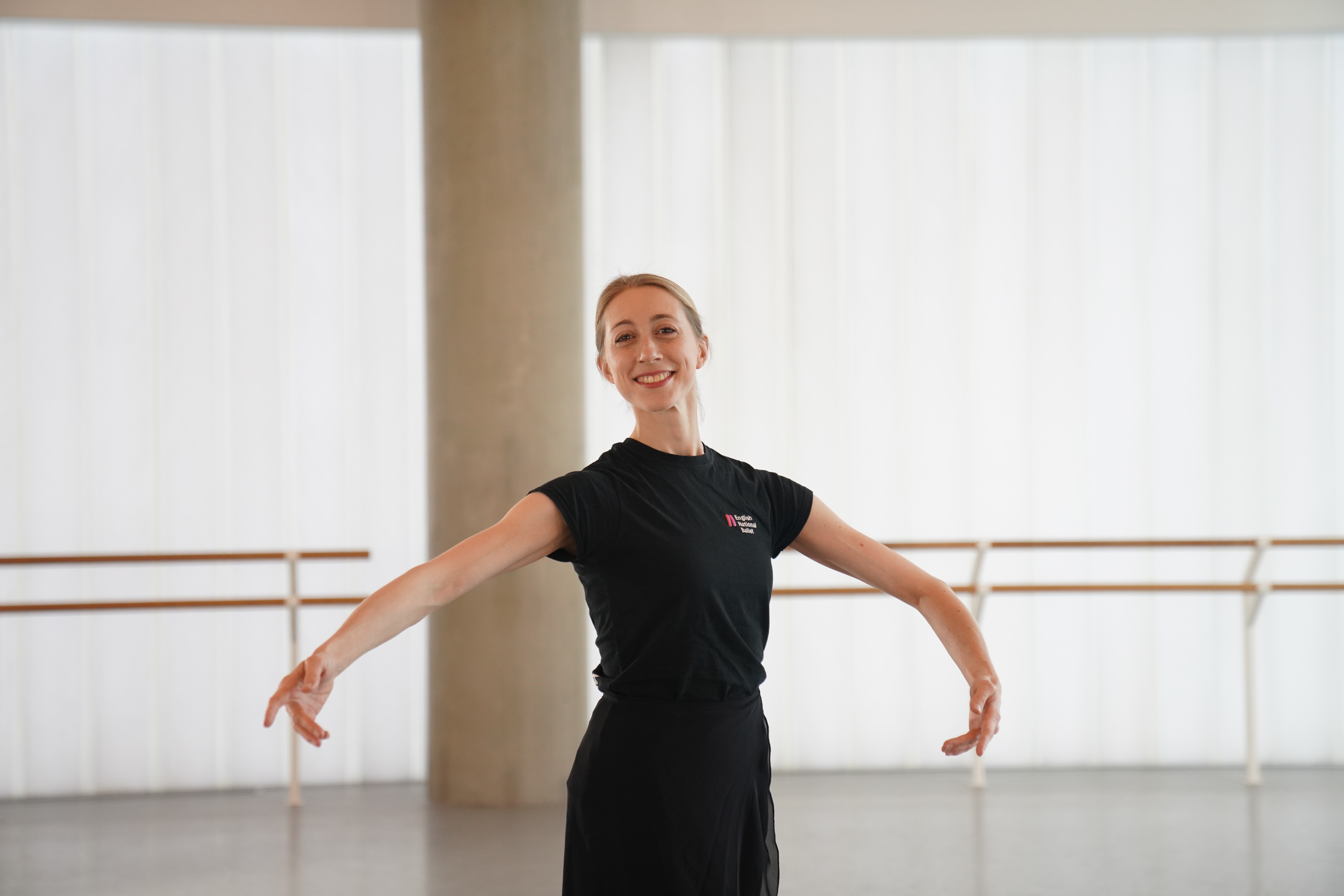 Ballet dancer smiling with arms open in a bright studio, wearing black and standing in front of ballet barres and tall windows.