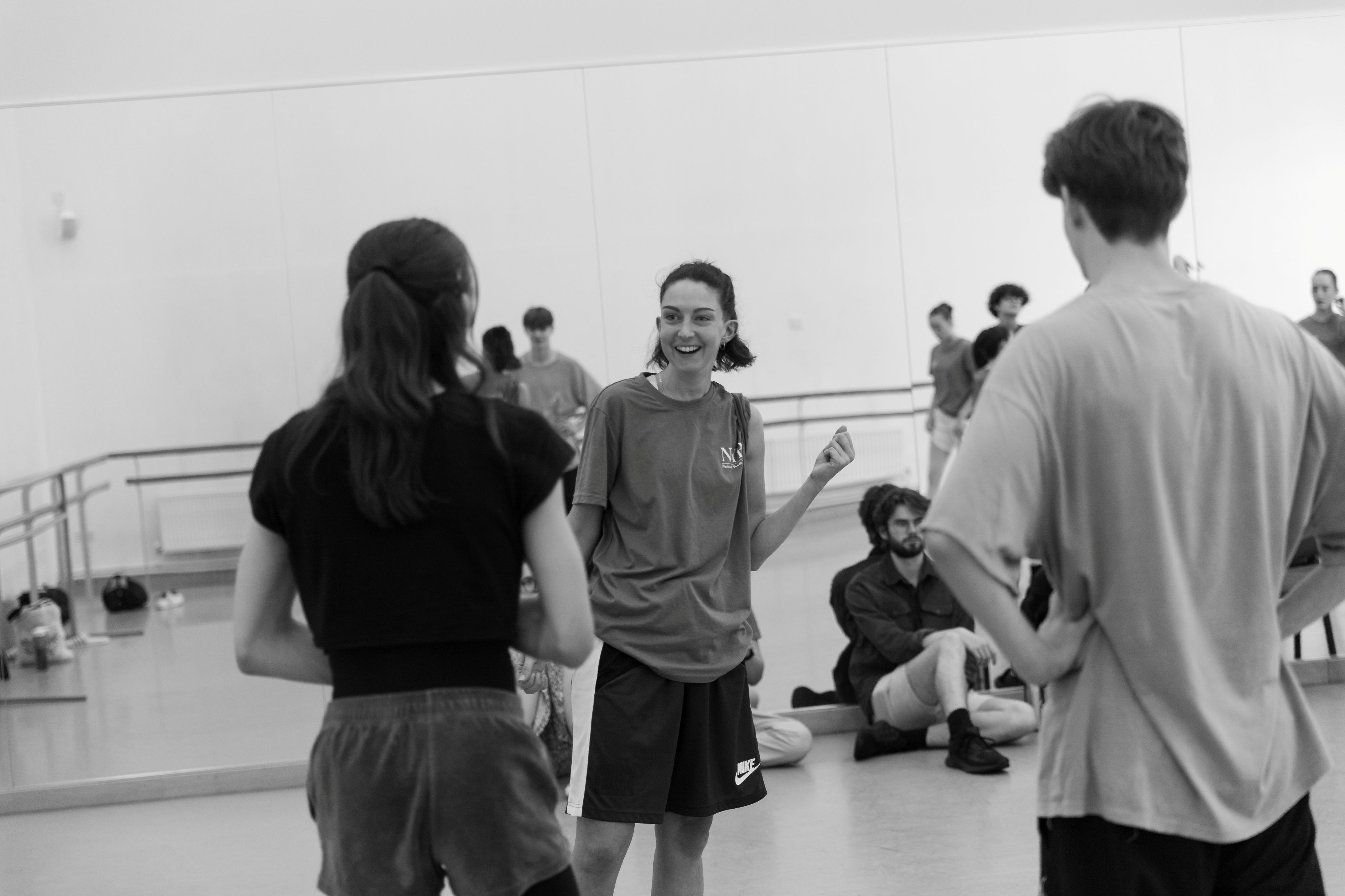 Smiling dance teacher stands in the middle of a dance studio talking with two people.