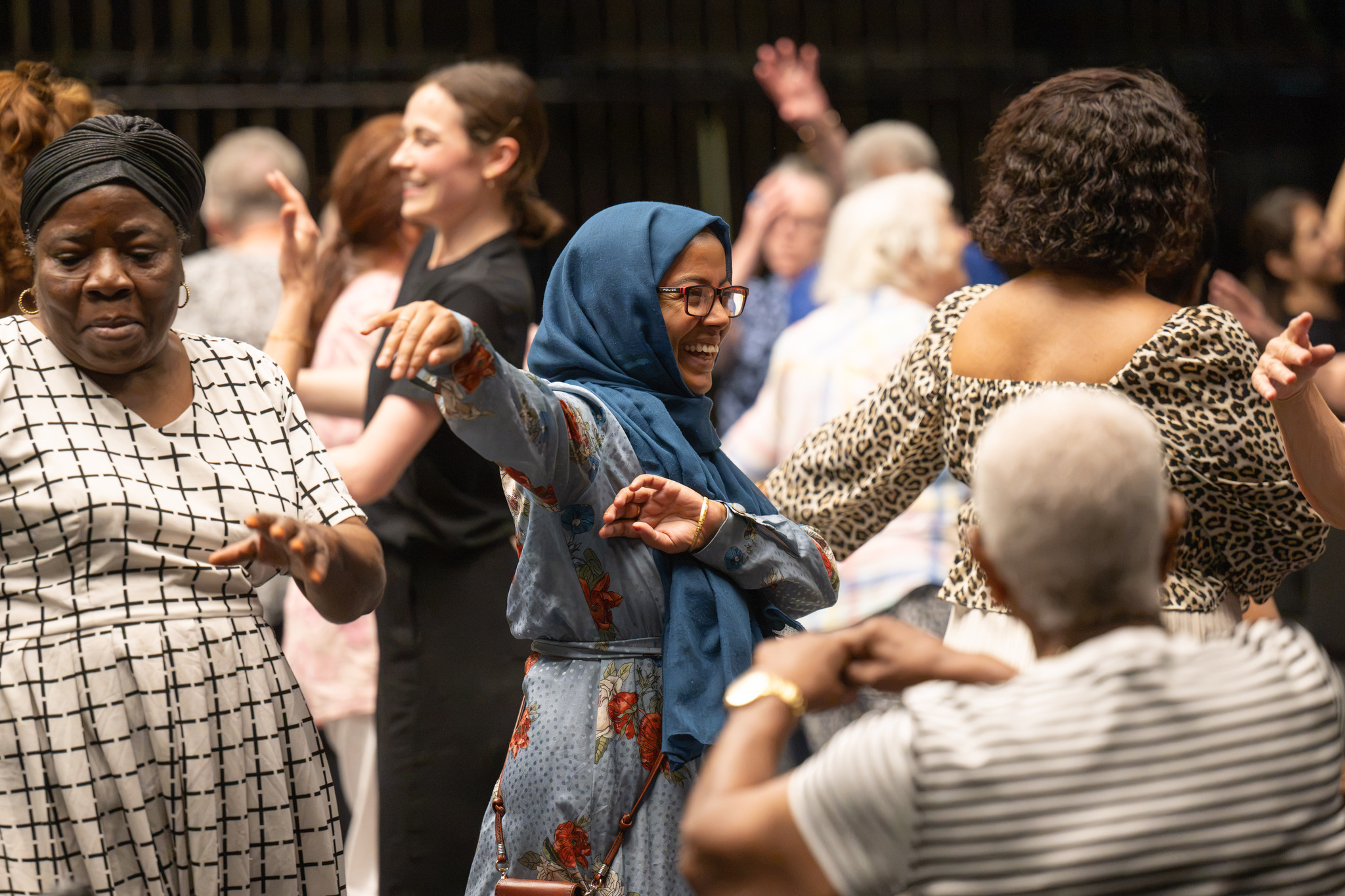 A group of adults smiling and moving together during a dance session, with participants raising and extending their arms. A woman in a blue headscarf and glasses is at the centre of the group with her arms outstretched.
